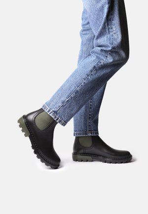 Jambes en jean bleu portant des bottes Chelsea noires avec des inserts élastiques verts, un pied levé, présentées sur un fond blanc.