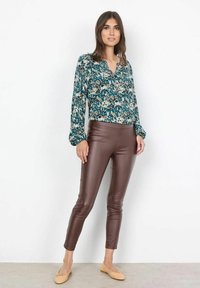 Blouse teal à imprimé floral avec manches longues, associée à un legging en faux cuir marron et des chaussures plates beiges, sur un fond neutre.