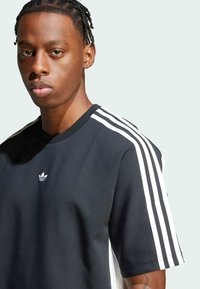 Melns īsām piedurknēm T-krekls ar apaļu kakla izgriezumu, uz kura pleciem ir baltas Adidas trīs svītru akcenti un neliels logo uz krūšu.