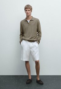 Beige poloko s dlouhým rukávem a límcem, ve spojení s bílými lněnými šortkami. Hnědé lodní boty dotvářejí celý outfit. Jednoduché pozadí.