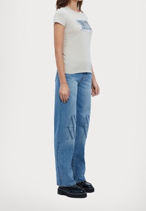 Kvinde iført hvid top med korte ærmer med sølvfarvet firkantet grafik og blå jeans med vide ben med stort “AX”-tryk foran på lårene.