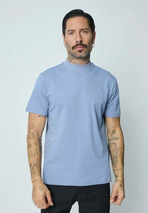 T-shirt bleu clair à manches courtes en tissu doux et lisse avec un design à col haut. Présente des tatouages visibles sur les deux bras.