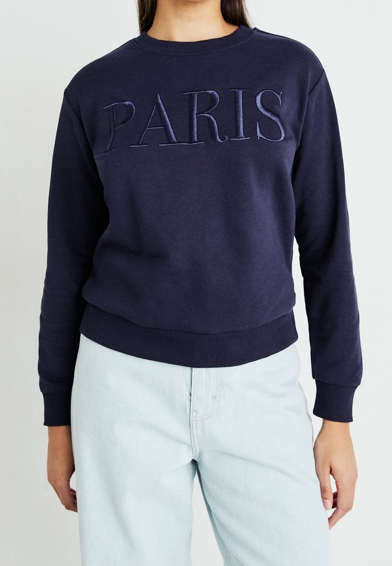 Personne portant un sweat-shirt bleu marine avec "PARIS" brodé sur la poitrine et un pantalon bleu clair sur un fond uni.