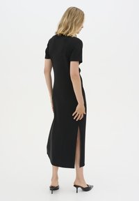 Black midi dress with short sleeves, fitted bodice, side slits, and smooth texture. Model wears black open-toed heels. Simple, elegant design.