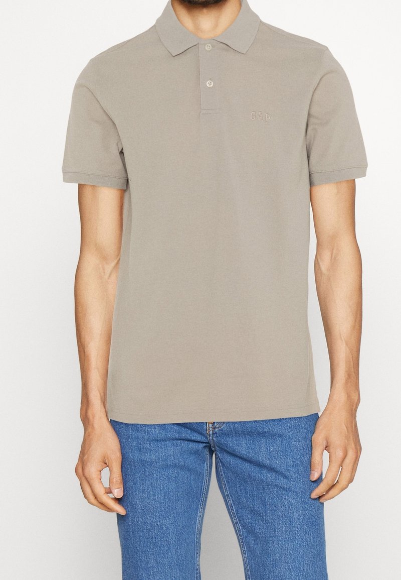 Homme portant un polo beige à manches courtes avec deux boutons et un jean bleu, debout devant un fond blanc uni.