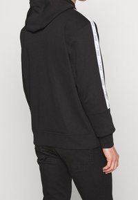 Svart hoodie med avslappnad passform, med vit Nike-logoband längs ärmarna. Mjuk tyg, justerbar dragsko huva, ribbad manschett.