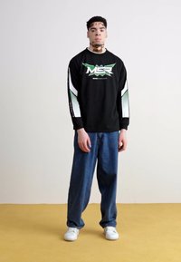 Schwarzes Langarmshirt mit grünen und weißen Akzenten und Logo, kombiniert mit lockeren blauen Jeans und weißen Sneakers auf einem gelben Boden.