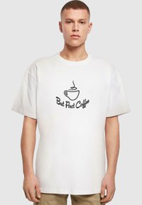 Hvid bomulds T-shirt med korte ærmer, prydet med et sort grafik af en dampende kaffekop og teksten "Men først kaffe" nedenfor.
