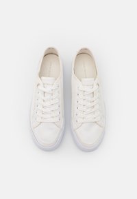 Valkoiset kangastennarit, joissa on teksturoidu pinta, pyöreä kärki ja valkoiset nauhat. Kielekkeessä on hillitty brändäys ja pohja on sileä ja valkoinen.