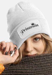 White knit beanie with a ribbed texture features a rectangular patch displaying the word "Prinzessin" in black cursive and a crown graphic.