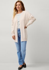 Cardigan rosa chiaro realizzato in morbido tessuto a maglia, design aperto sulla parte anteriore, con polsini a coste e vestibilità rilassata, abbinato a jeans azzurri.
