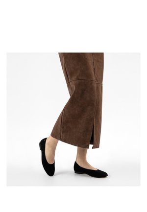 Nohy v hnědé semišové sukni s bočním rozparkem a černých balerínkách, stojící na bílém pozadí.
