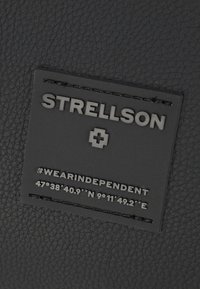 Etichetta in pelle nera texturizzata con la scritta "STRELLSON", una croce svizzera e coordinate con l'hashtag #WEARINDEPENDENT. Finitura in rilievo e opaca.