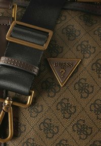 Dettaglio di una borsa in tessuto marrone con motivo, con logo triangolare dorato, tracolla nera con fibbia e dettagli in metallo dorato.