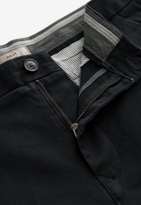 Pantalon noir avec fermeture éclair partiellement ouverte, taille boutonnée et détail visible de la doublure intérieure rayée.