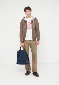 Καφέ φούτερ με φερμουάρ και γκρι επένδυση, λευκό μπλουζάκι με γραφικό, ανοιχτό καφέ παντελόνι και μπλε τσάντα tote από τζιν με δερμάτινες λεπτομέρειες. Καφέ παπούτσια.