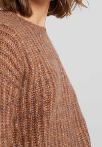 Pull en tricot marron avec une texture côtelée, un col large et une coupe décontractée. Le matériau semble doux et chaud.