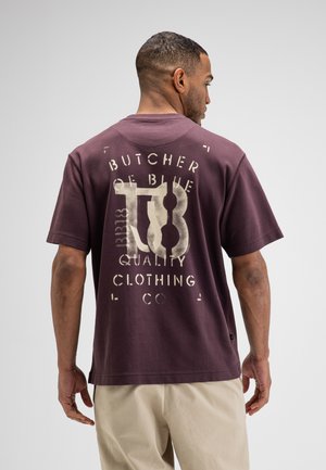 Man draagt een donkerpaarse T-shirt met grote witte tekst "Butcher of Blue TB Quality Clothing" op de rug, gecombineerd met beige broek.