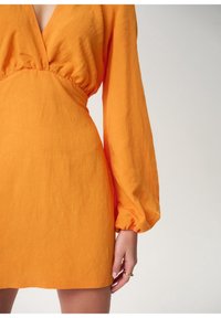 Oranžové šaty s dlouhým rukávem a hlubokým výstřihem do V, se skládáním v pase. Materiál vypadá lehce a má jemný strukturovaný povrch.