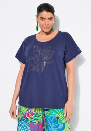 T-shirt blu navy a maniche corte, con un design floreale metallico. Abbinato a pantaloni colorati e decorati che raffigurano foglie tropicali.
