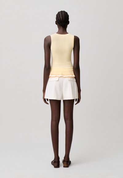 Mujer con el pelo trenzado que lleva una blusa sin mangas de color amarillo pálido con dobladillo con volantes, pantalones cortos blancos con bolsillos y sandalias oscuras, de espaldas.