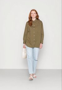 Camisa verde-oliva de botão, com mangas longas e um único bolso no peito, combinada com jeans rasgados de tom azul claro e sandálias claras.