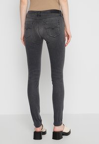 Femme portant un jean skinny noir avec poches arrière et des mules beiges à talons, debout devant un fond gris clair uni.