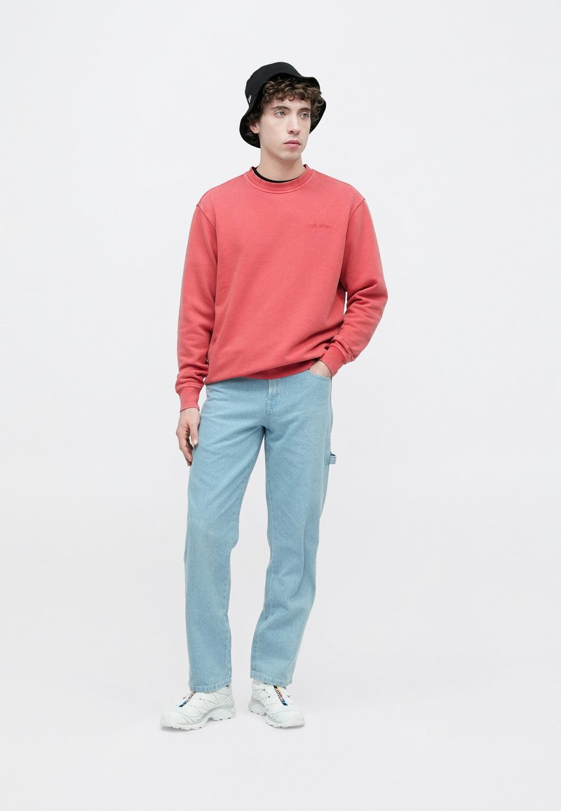 Felpa rossa con scollo a girocollo, jeans azzurri e sneakers bianche. Cappello nero a secchiello. Outfit casual con dettagli minimi e vestibilità rilassata.