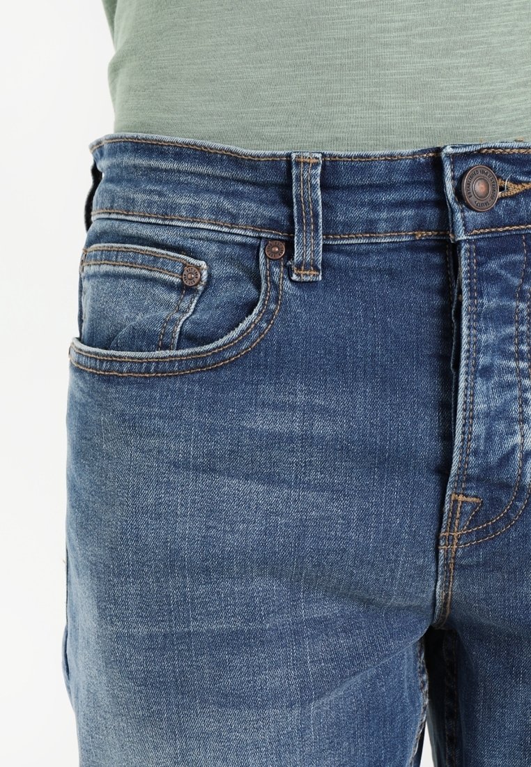 Gros plan sur le torse portant un jean en denim bleu et une chemise vert clair rentrée, montrant le bouton, les passants de ceinture et les poches.