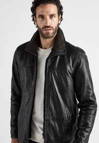 Schwarze Lederjacke mit Reißverschluss, strukturiertem braunem Kragen, schlichtem Design, seitlichen Taschen und leicht taillierter Silhouette.