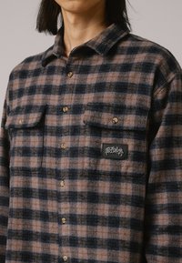 Camicia di flanella con motivo a quadri marrone e blu navale. Presenta due tasche sul petto con patte e una toppa con logo. Tessuto strutturato con colletto.