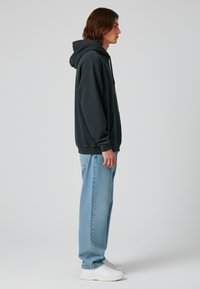 Felpa oversize grigio scuro con tasca frontale e cappuccio con coulisse, abbinata a jeans blu chiaro a vestibilità comoda e sneaker bianche.