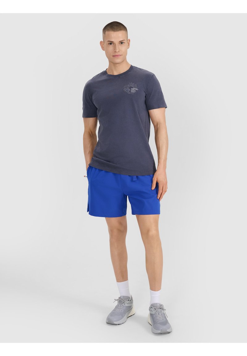 Jeune homme debout portant un t-shirt gris foncé, un short bleu vif, des chaussettes blanches et des baskets grises sur un fond clair uni.