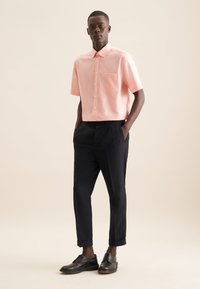 Camicia rosa a maniche corte con bottoni e tasca sul petto, abbinata a pantaloni neri aderenti e scarpe nere stringate.