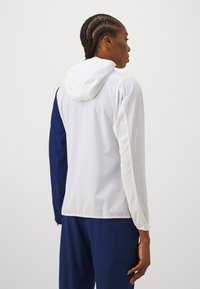 Viegls kapuces jaka ar baltu korpusu un tumši zilu piedurknēm, izgatavota no gludas auduma ar elastīgām piedurknēm ciešai piegulšanai.