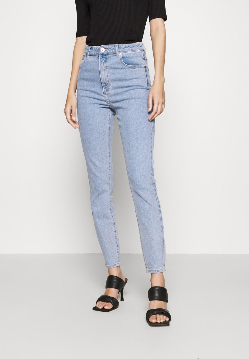 Vaaleansiniset korkeavyötäröiset skinny-farkut, joissa on vetoketjukiinnitys ja nappisulku. Yhdistetty mustiin avonaisiin korkokenkiin. Sileä denim-pinta.