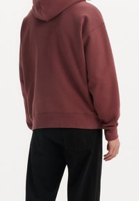 Osoba v bordovém mikině s kapucí spuštěnou a černých kalhotách, zobrazená zezadu na jednoduchém bílém pozadí.