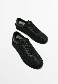 Musta matala malli tennari, joissa on verkkokyljet ja nahkakerrokset, varustettuna mustilla kengännauhoilla valkoista taustaa vasten.