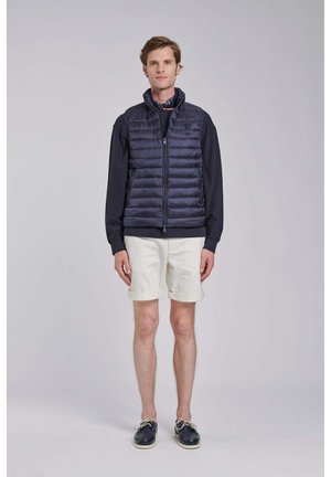 Mężczyzna noszący granatową kamizelkę puchową na czarnej koszulce z długim rękawem, białe podwinięte szorty, granatowe mokasyny oraz chustę na szyi, stojący na jednolitym tle.