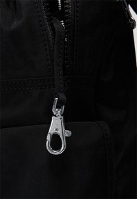 Primer plano de una mochila de tejido negro, con un tirador de cremallera plateado y un clip. El material tiene un acabado texturizado y costuras reforzadas.