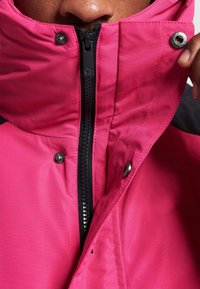 Fuchsia jakke med høy krage, med svart glidelås og sølvfarge trykknapper. Stoffet fremstår som glatt og værbestandig.