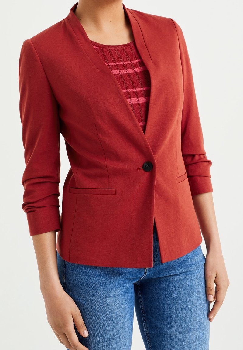 Blazer rouge ajusté avec un col cranté, une fermeture à un bouton et deux poches avant. Porté par-dessus un haut à rayures et un jean bleu.