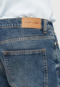 Τζιν τζιν με ξεβαμμένο μπλε φινίρισμα που διαθέτει καφέ δερμάτινη ετικέτα με την ανάγλυφη επιγραφή "SAM-SØE", αντίθεση ραφών και κανονικές πίσω τσέπες.