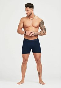 Boxer briefs blu navy con una cintura morbida, realizzati in tessuto elasticizzato; indossati da un modello maschile con tatuaggi e un fisico muscoloso.