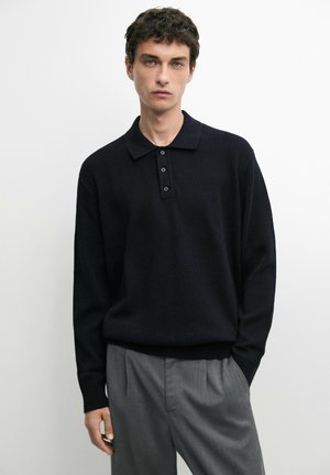 Giovane uomo con capelli corti e ricci che indossa una polo nera a maniche lunghe in maglia e pantaloni grigi su sfondo bianco semplice.