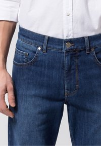 Jeans de mezclilla azul oscuro con corte recto, que cuentan con cinco bolsillos, herrajes plateados y detalles de costura en naranja contrastante.