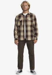 Bruine en beige geruite flanellen shirt met twee borstzakken, gecombineerd met groene broek en zwarte sneakers. Gewone witte t-shirt eronder.