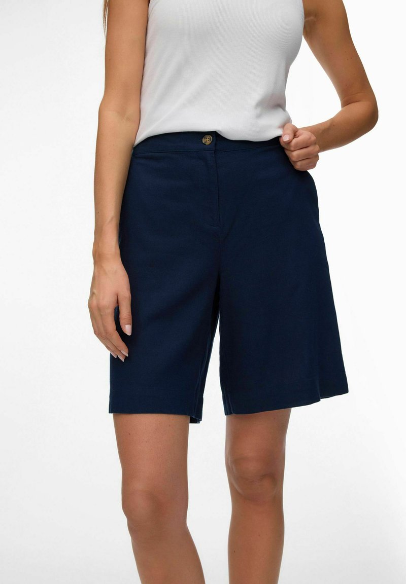 Femme portant un haut blanc sans manches et un short bleu marine ajusté arrivant aux genoux, debout, une main ajustant la taille.