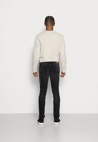 Man with braided hair wearing a beige sweatshirt, black jeans, and black sneakers standing facing a white wall.