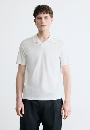 Jonge man met krullend haar, gekleed in een wit poloshirt met korte mouwen en een zwarte broek, staand tegen een eenvoudige lichte achtergrond.